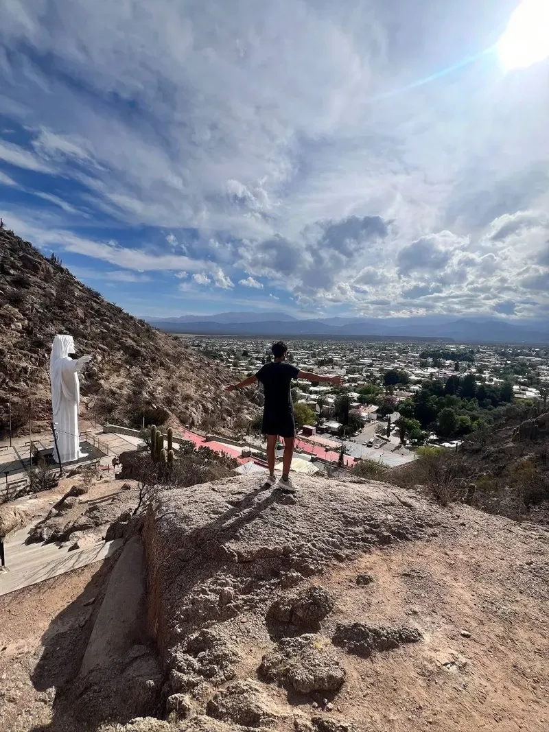 Sergi con los brazos abiertos en el mirador del Cristo del Portezuelo, con la estatua blanca a la izquierda y la ciudad de Chilecito abajo