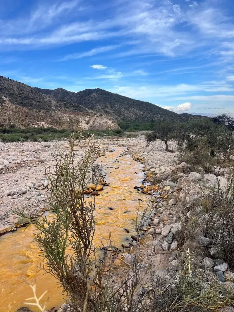 Río de agua naranja entre cantos rodados con montes y cielo azul al fondo