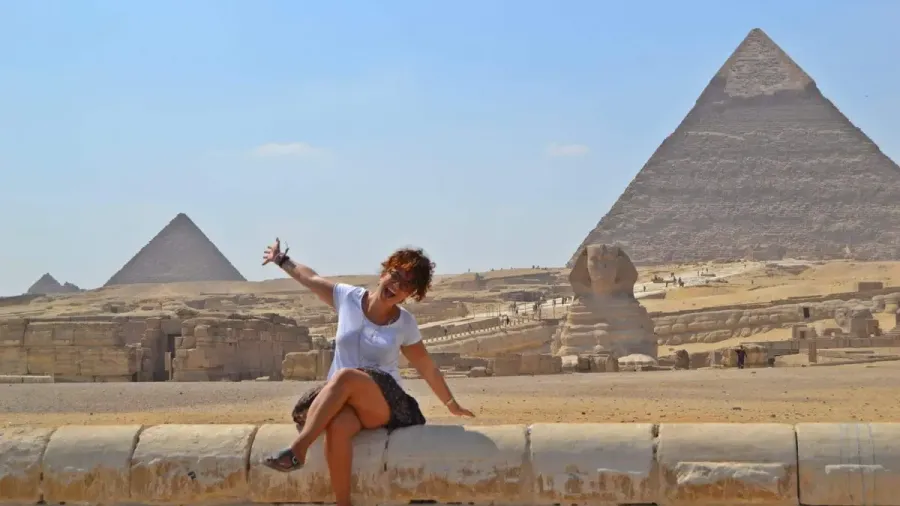 Mujer sentada en un muro posando con los brazos abiertos frente a las pirámides de Giza