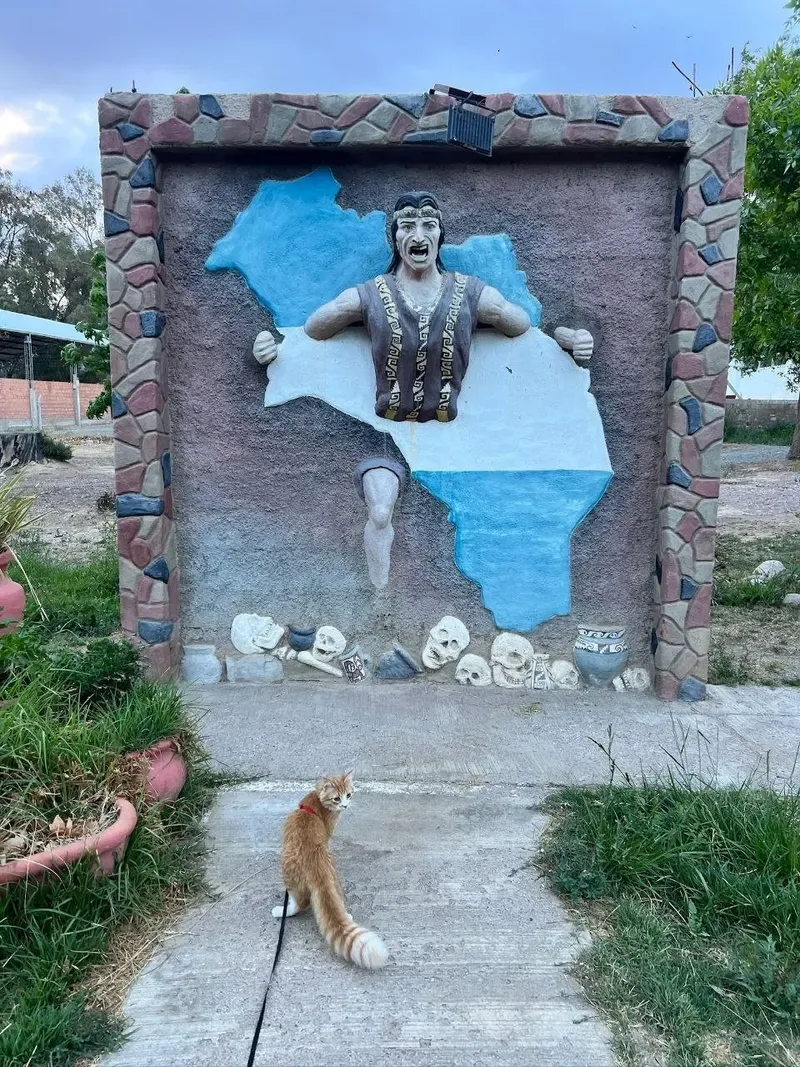 Mural de un indígena que rompe el mapa de La Rioja, con cráneos y vasijas, y un gato naranja con arnés delante