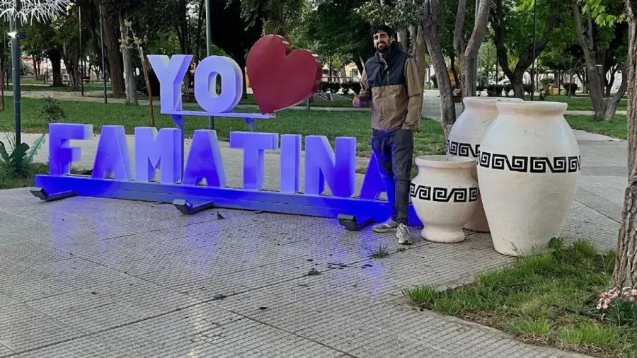 Sergi junto al cartel luminoso «Yo corazón Famatina» y tres vasijas blancas con grecas negras, en un parque del pueblo