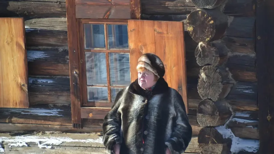 Mujer mayor con abrigo de piel junto a una casa de madera tradicional siberiana