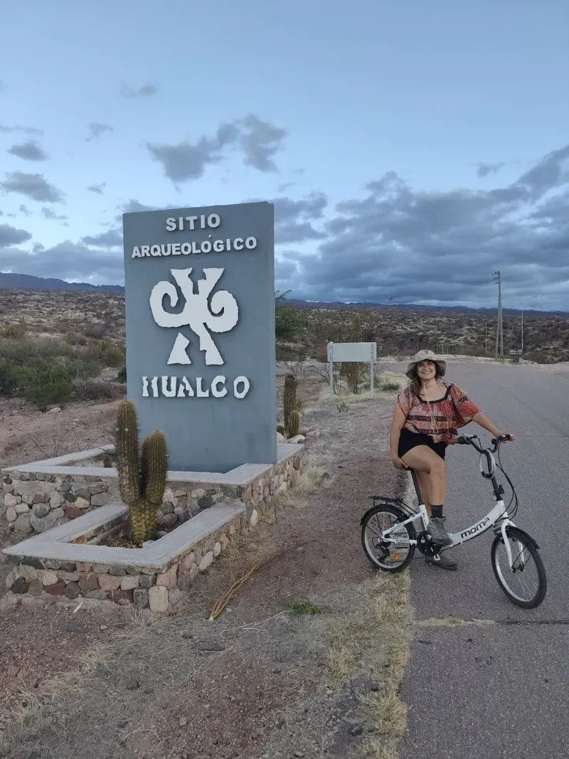 Cris con una bicicleta plegable junto al cartel del Sitio Arqueológico Hualco, en el borde de la carretera