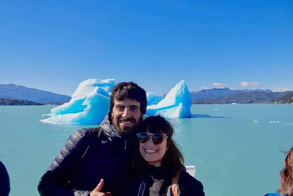 Sergio y Cris sonriendo a bordo de un barco con un témpano de hielo de fondo en el Lago Argentino
