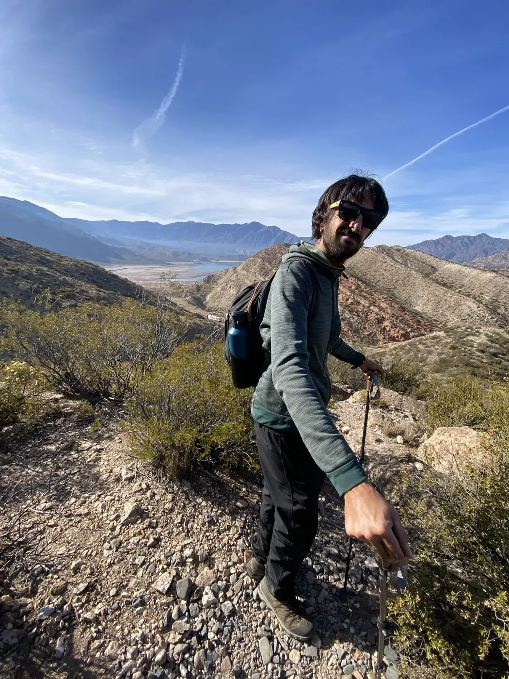 Un viajero con bastones de trekking en un sendero sobre el embalse de Potrerillos, con montañas y el lago al fondo