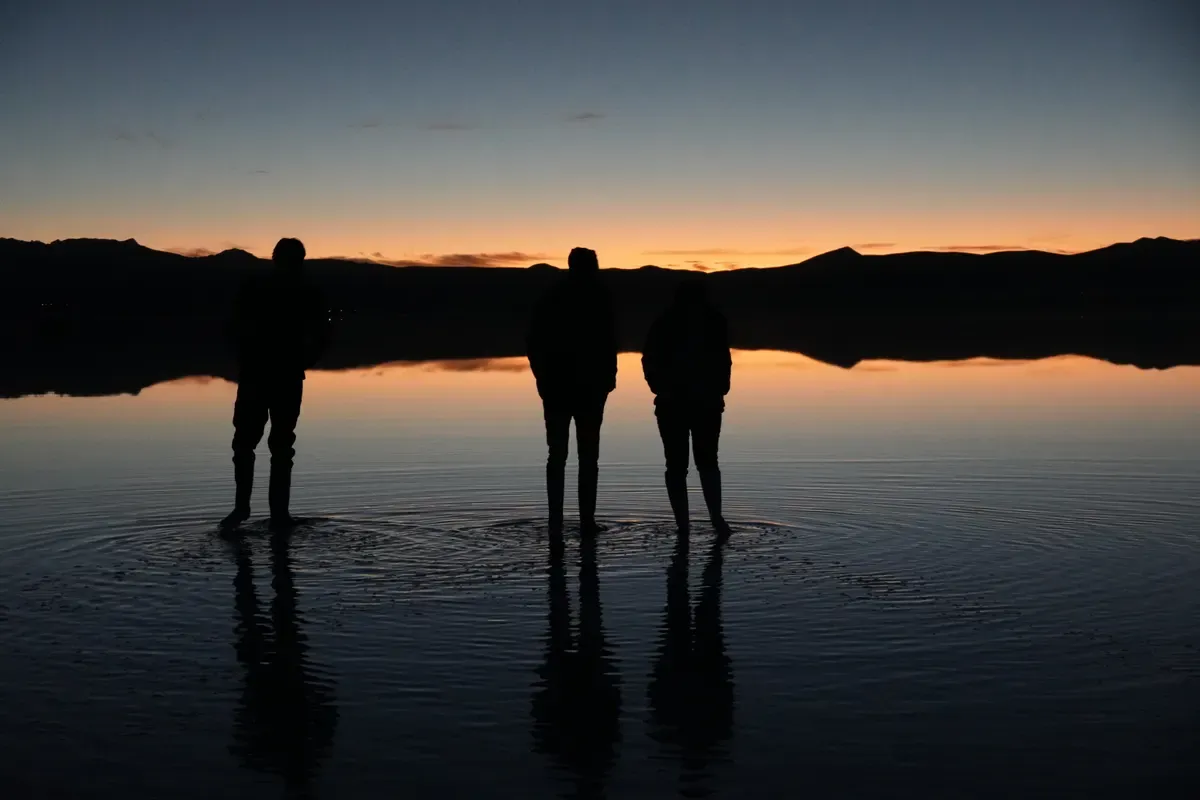 Tres siluetas de pie sobre el agua del Salar de Uyuni al amanecer, con el cielo naranja reflejado como un espejo