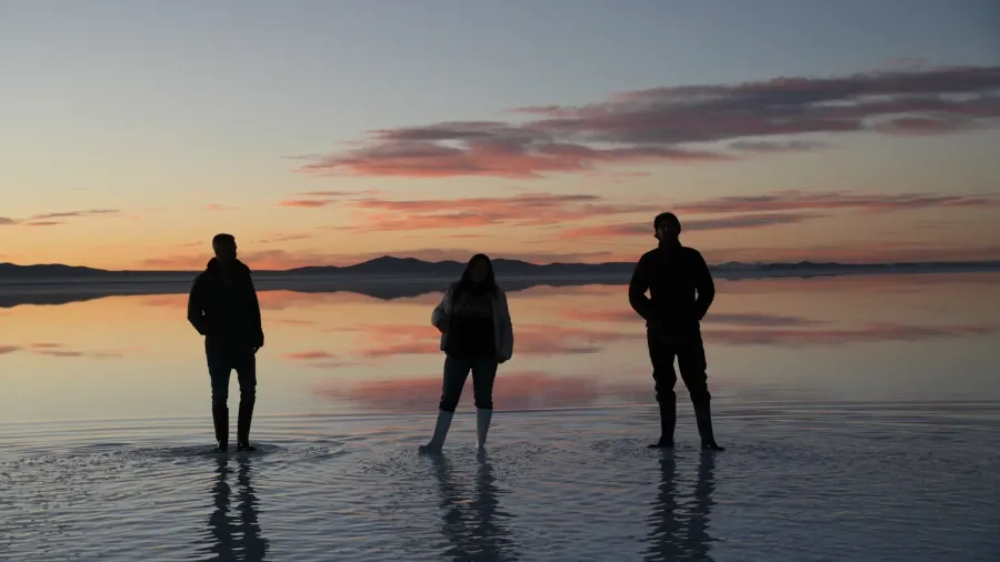 Tres viajeros de pie sobre el agua del Salar de Uyuni al amanecer, con las nubes rosas y naranjas reflejadas como un espejo