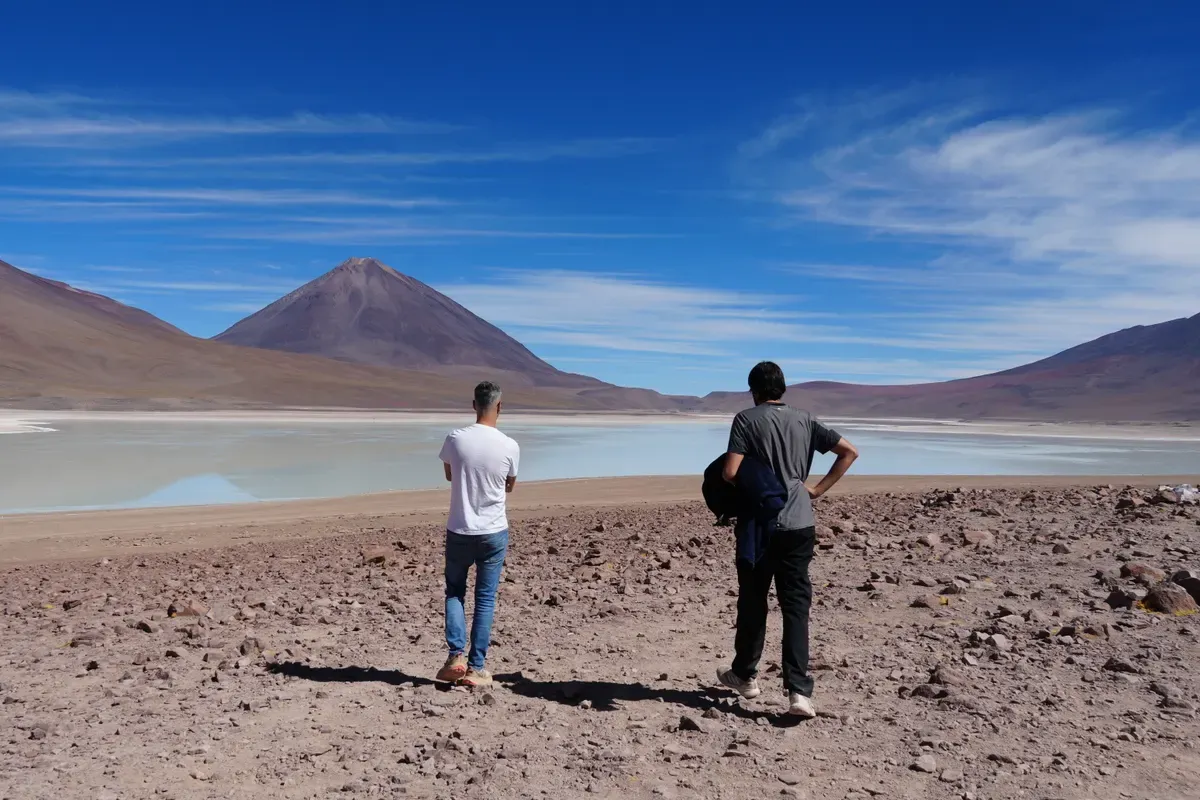 Dos viajeros frente a una laguna del altiplano con un volcán y el cielo azul al fondo