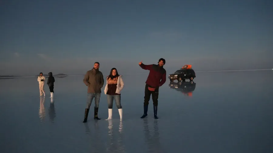 Tres viajeros posando con chaquetas y botas altas sobre el Salar de Uyuni inundado antes del amanecer, con un todoterreno al fondo