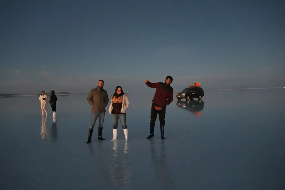Tres viajeros posando sobre el Salar de Uyuni inundado antes del amanecer, con un todoterreno reflejado en el agua al fondo