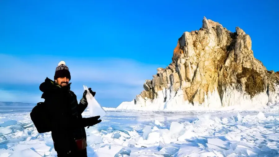 Viajero abrigado sosteniendo un trozo de hielo frente a la roca Shamanka en el lago Baikal helado
