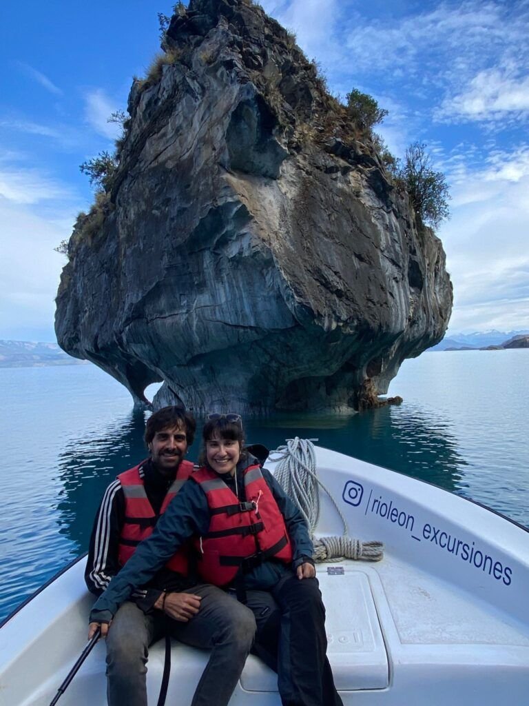 Cris y Sergi con chalecos salvavidas rojos sentadas en una lancha frente a una gran formación rocosa sobre aguas turquesa en un lago rodeado de montañas.