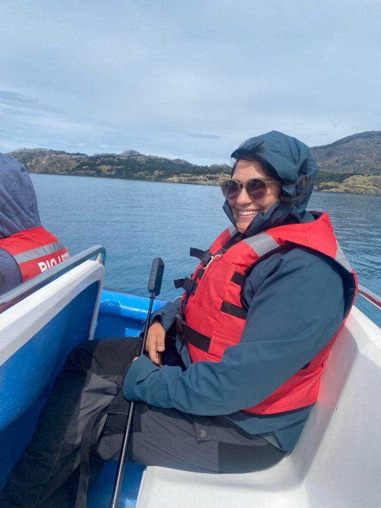 Cris sonriente con chaleco salvavidas rojo y gafas de sol navegando en una pequeña embarcación sobre un lago rodeado de montañas.