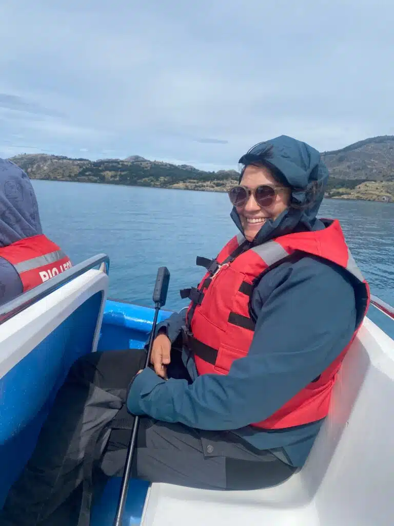 Cris sonriente con chaleco salvavidas rojo y gafas de sol navegando en una pequeña embarcación sobre un lago rodeado de montañas.