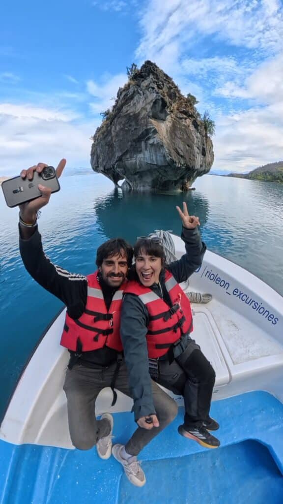 Pareja con chalecos salvavidas haciéndose un selfie en una lancha frente a la Capilla de Mármol, formaciones rocosas sobre aguas turquesas en la Patagonia chilena.